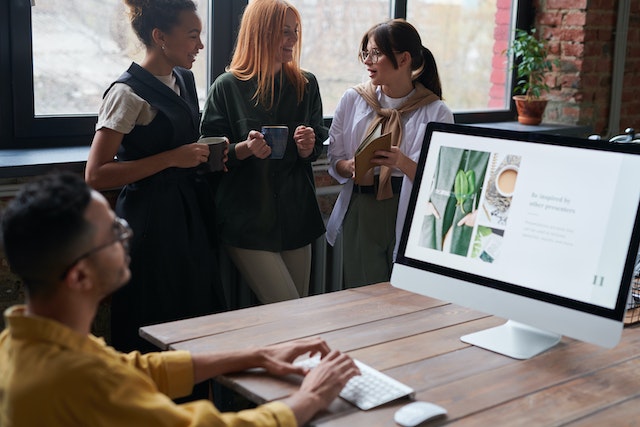 Three businesswomen casually have a discussion holding coffee mugs by a window while a man sits at his hot desk working on a PC at a hot desking and virtual office in London image at LondonOfficeSpace.com.