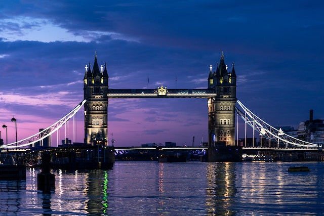 Dusk view down the River Thames of the illuminated Tower Bridge spanning the river with a purple and blue cloud-strewn sky in the background and riverbanks lined with buildings with postcodes in Central London.