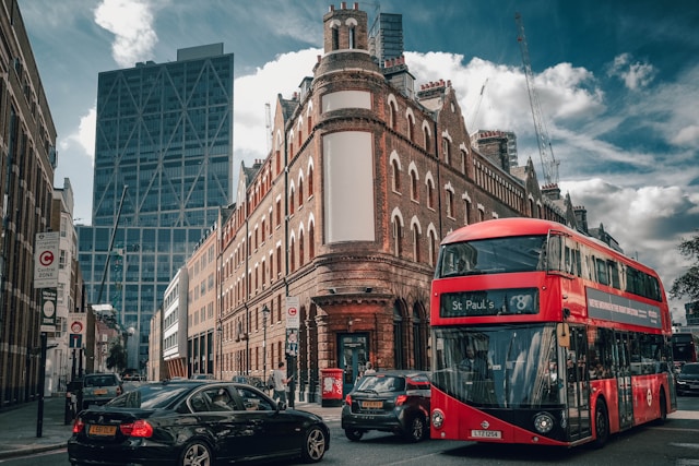 A daytime view at the apex of a busy street junction in Mayfair with many cars driving by, including one of London’s iconic red double-decker buses. The streets border a historic wedge-shaped building running down the centre, and there’s a modern skyscraper in the background framed by a cloud-strewn sky. Image at LondonOfficeSpace.com.