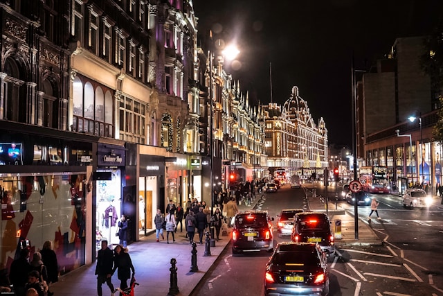 Night view down a street in Fitzrovia. Shopfront lights illuminate people walking on the sidewalks, and cars pack the street, including London’s iconic black cabs. Image at LondonOfficeSpace.com.
