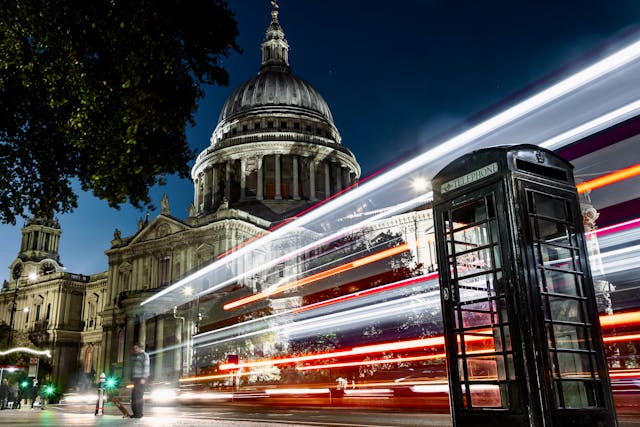 A night view of St. Paul’s Cathedral from the side walk next to one of London’s iconic red telephone boxes, and a double-decker bus is streaking past on the street between the phone box and the cathedral. Image at LondonOfficeSpace.com.