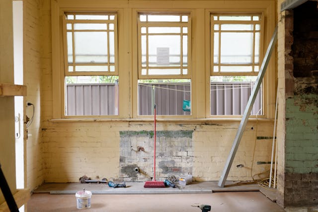 A view from inside of a yellow wall inset with three windows that is being remodeled. A red broom leans on the middle of the windows' shared awning and various other building supplies litter the floor around the wall. Image at LondonOfficeSpace.com.