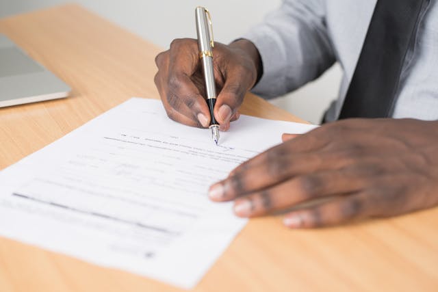A close-up view of a man in a business shirt and tie signing a document with a silver pen resting on a light wood desk. Image at LondonOfficeSpace.com.