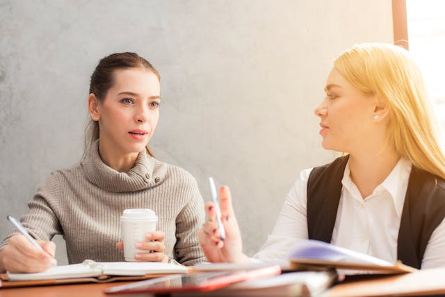 Two women sitting at a desk negotiating terms. They each hold pens and have notebooks in front of them. Image at LondonOfficeSpace.com.