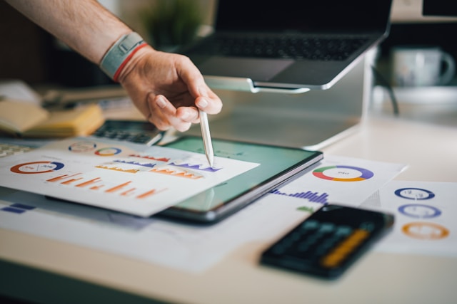 A hand holding a pen points to printed cost comparison graphs resting on a tablet on a desk with other printed graphs, a calculator, and a laptop also littering the desk. Image at LondonOfficeSpace.com.