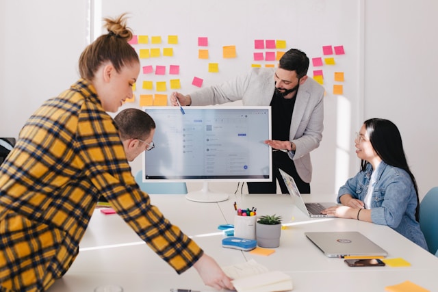 A team of colleagues, two men and two women, sit and stand around a desk in a bright, white meeting room. A male in a grey jacket points at a screen at the head of the desk and there's a whiteboard with yellow, pink, and orange post-it notes stuck to it behind the man. Image at LondonOfficeSpace.com.