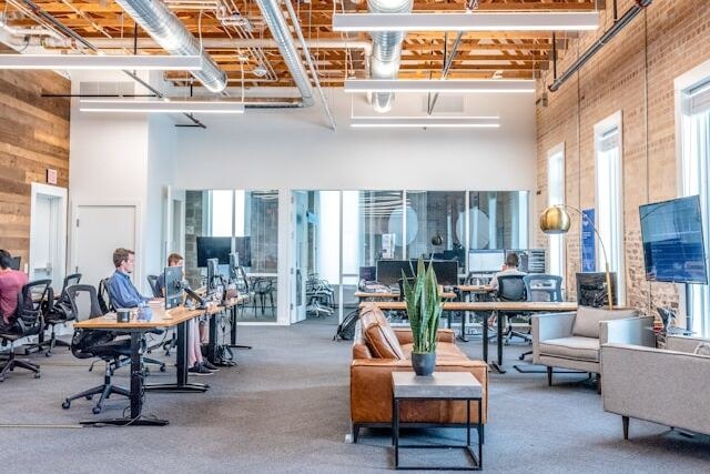 A view across a modern, well-lit, and high-ceilinged workspace. There is exposed, shiny ducting criss-crossing the ceiling as well as bright, straight light fixtures hanging down. People work at desks on the left, there's breakout space on the right next to tall windows, and there's a private office and meeting room behind glass on the far wall. Image at LondonOfficeSpace.com.