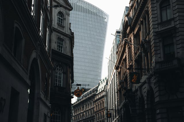 A street view from between multi-storey, old buildings up towards the famous 20 Fenchurch Street skyscraper. The sky is mostly blocked out by the skyscraper, but what little there is visible is overcast and grey. The old buildings that rise up on all sides of the street are mostly shrouded in shadow. Image at LondonOfficeSpace.com.