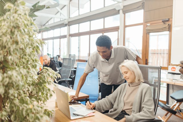 A smiling woman with white hair and wearing a loose grey cardigan is seated at a desk in a coworking facility brightly backlit by sunlight from a peaked wall of windows with her laptop in front of her. She’s holding a pen and writing in a notebook that rests on her laptop, while a smiling man in a grey polo shirt standing to her side is leaning over her and explaining something. Image at LondonOfficeSpace.com.