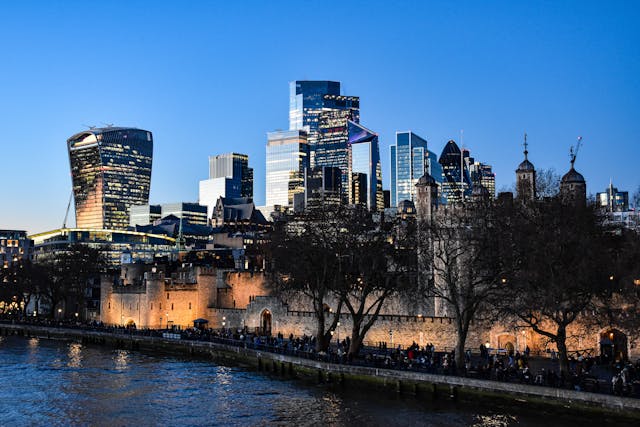 A dusk view from over the River Thames towards the Tower of London, whose ancient walls are lit up with orange lights, with the gleaming skyscrapers of modern London in the background. Many of the skyscrapers’ windows are lit up with interior lights, and the glass facades of a few buildings reflect the setting sun under a blue, cloudless sky. Image at LondonOfficeSpace.com.