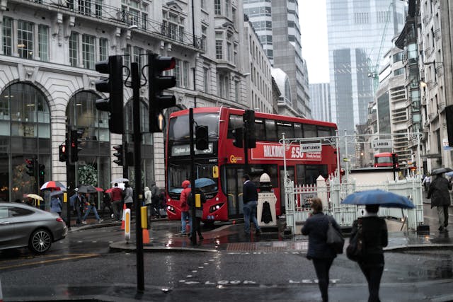 Daytime view from a footpath looking towards an intersection in central London. Historic buildings line the street and there are gleaming skyscrapers in the distance with a grey, overcast sky peeking out between them. The road is shiny with rain and most people walking along the street are carrying umbrellas. One of London’s iconic red buses is about to pass through the intersection, while people wait to cross the cross-walk on foot. Image at LondonOfficeSpace.com.