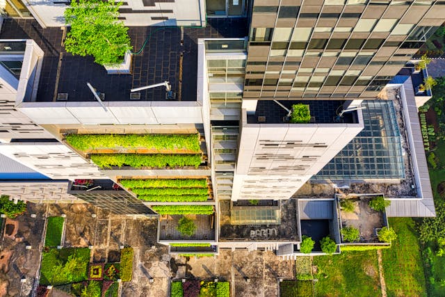 A drone’s view down the length of a tall building’s facade. The balconies that run up most levels of the building on the left are packed with hanging green plants, at the base of the building on the right, there is a large greenhouse, and cultivated gardens surround the rest of the building’s base. Greenhouses and cultivated grounds to grow fruits and vegetables, as well as abundant green spaces for building occupants, are great ways to improve a building’s BREEAM rating. Image at LondonOfficeSpace.com.