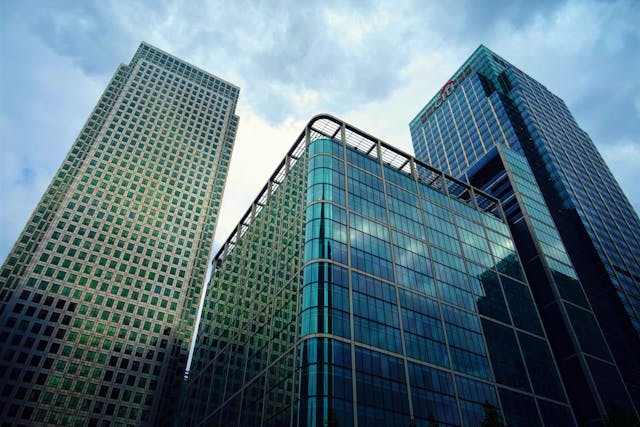 A low-angle daytime view up at three towering skyscrapers in the London business district. The buildings are modern with glass facades shining with a turquoise hue under a cloudy sky. Modern London office buildings are leading the way when it comes to sustainable office design, many attaining Excellent or Outstanding BREEAM ratings. Image at LondonOfficeSpace.com.