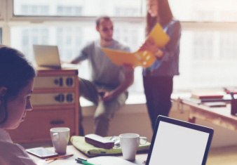 women on laptop with two colleagues standing and meeting in the background